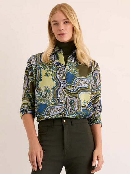 A woman stands against a plain background, wearing the Hinson Wu Margot Long Sleeve Opulent Paisley Shirt in Olive Paisley—crafted from Italian polyester fabric with a fluid drape and a silky hand feel—with the sleeves rolled up, layered over a dark turtleneck and dark pants.