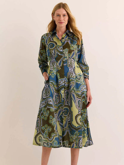 A woman stands against a plain light background, wearing the Hinson Wu Martina 3/4 Sleeve Opulent Dress in Olive Paisley—a button-up midi dress featuring a green, blue, and yellow paisley pattern, expertly crafted from Italian polyester fabric for a fluid drape and silky hand feel.