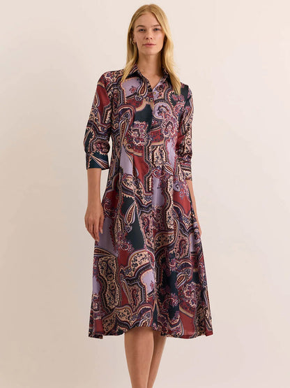 A woman stands against a plain light background with hands at her sides, wearing the Hinson Wu Martina 3/4 Sleeve Opulent Dress in Plum Paisley—a collared midi dress featuring a paisley pattern and crafted from Italian polyester fabric that drapes elegantly over the silhouette.