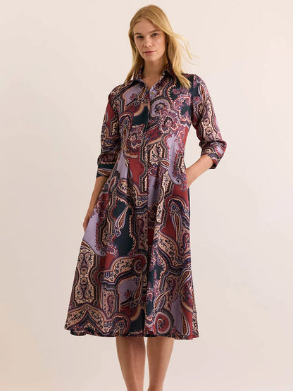 A woman stands against a plain light background, wearing the Hinson Wu Martina 3/4 Sleeve Opulent Dress in Plum Paisley—a midi-length, button-down dress with pockets, featuring an elegant paisley pattern and crafted from Italian polyester fabric for a silky feel and fluid drape.