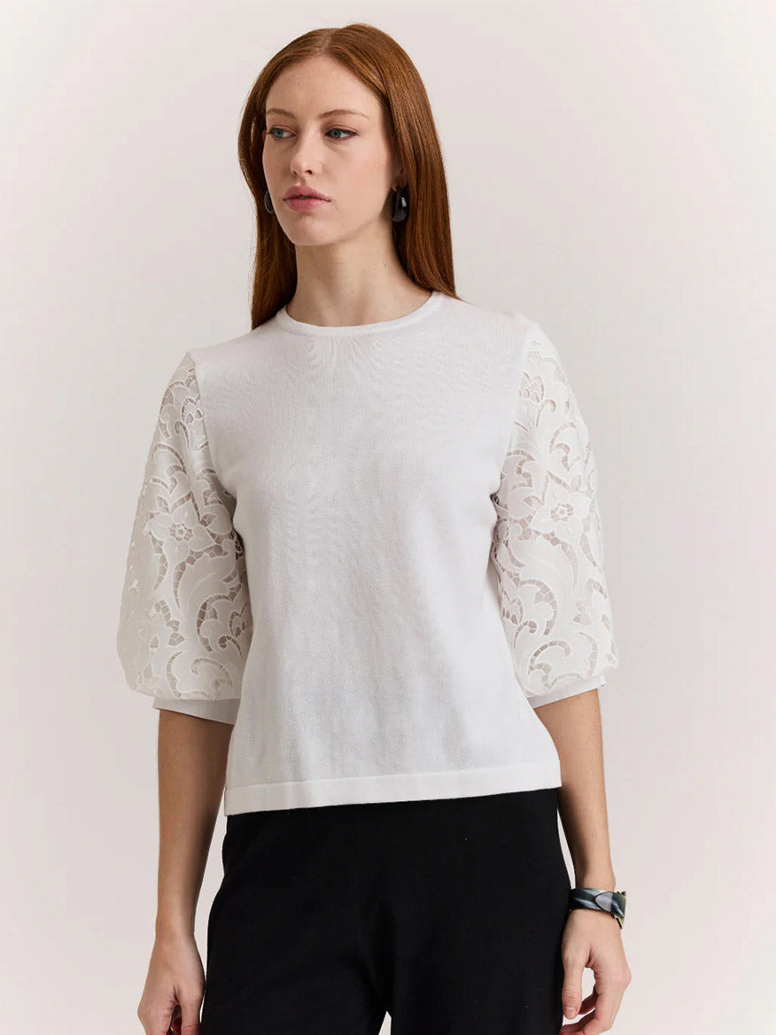 Woman wears the Hinson Wu Millie Lace Elbow Sleeve Sweater in White, featuring lace puff sleeves and floral patterns, paired with black pants—a chic look against a light background.