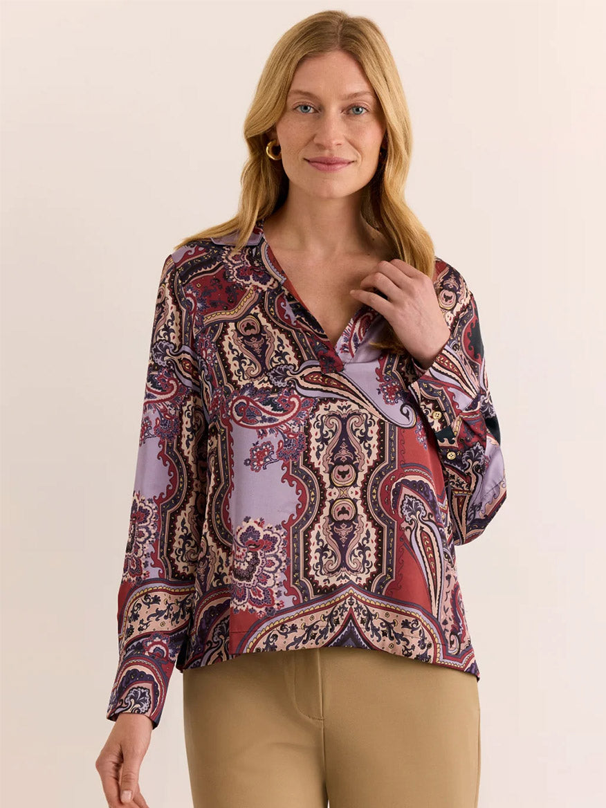 A woman with blonde hair stands against a plain light background, wearing the Hinson Wu Mona Long Sleeve Opulent Paisley Top in Plum Paisley—crafted from Italian polyester fabric with a silky hand feel in red, purple, and beige tones—and tan pants.