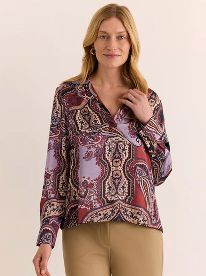 A woman with blonde hair stands against a plain light background, wearing the Hinson Wu Mona Long Sleeve Opulent Paisley Top in Plum Paisley—crafted from Italian polyester fabric with a silky hand feel in red, purple, and beige tones—and tan pants.