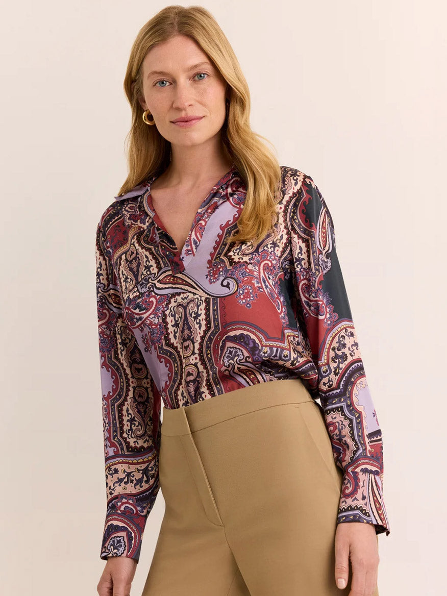 A woman stands against a plain light-colored background, wearing the Hinson Wu Mona Long Sleeve Opulent Paisley Top in Plum Paisley—crafted from Italian polyester with a fluid drape and silky hand feel in rich red, lavender, and black hues—paired with high-waisted tan trousers.