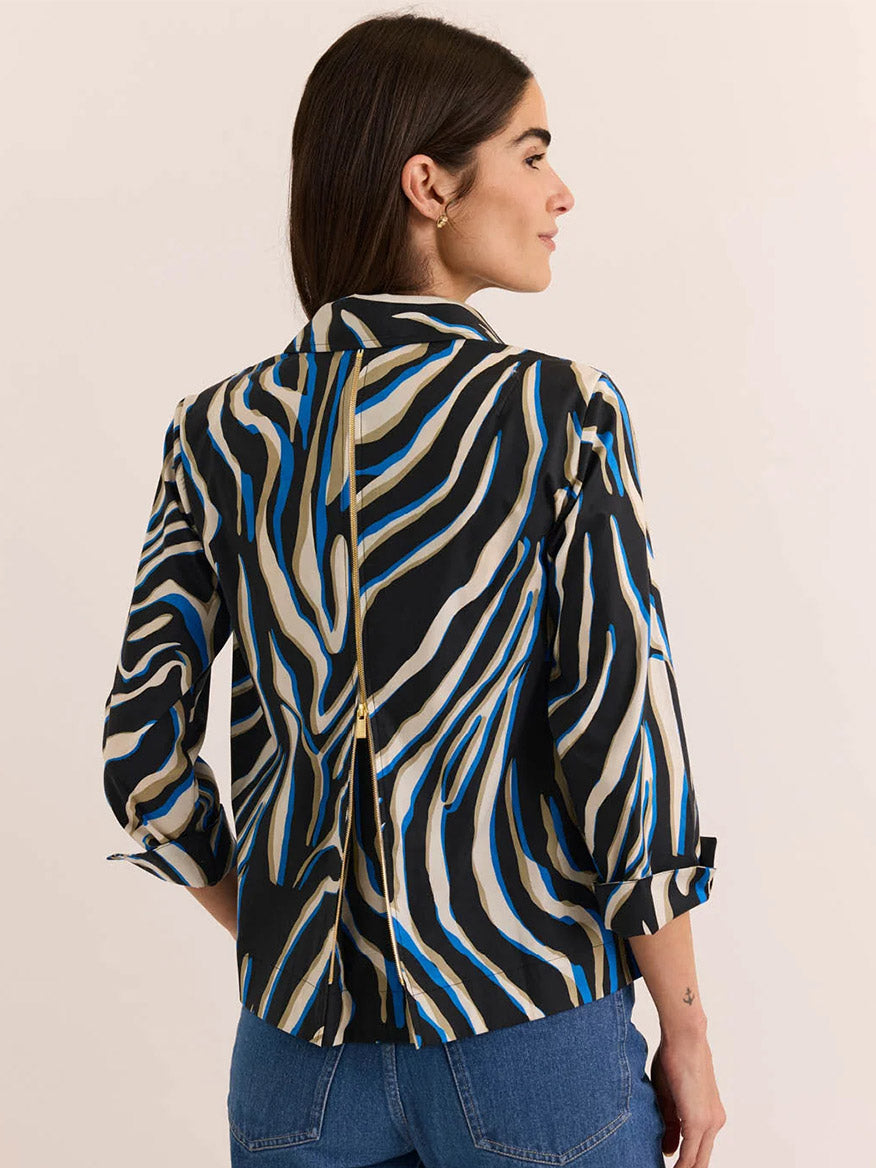 A woman facing away from the camera wears the Hinson Wu New Xena 3/4 Sleeve Shirt in Abstract Zebra—a button-down crafted from a premium cotton blend, showcasing a black, beige, and blue zebra print with a gold-colored zipper running vertically down the back. She pairs it with blue jeans.