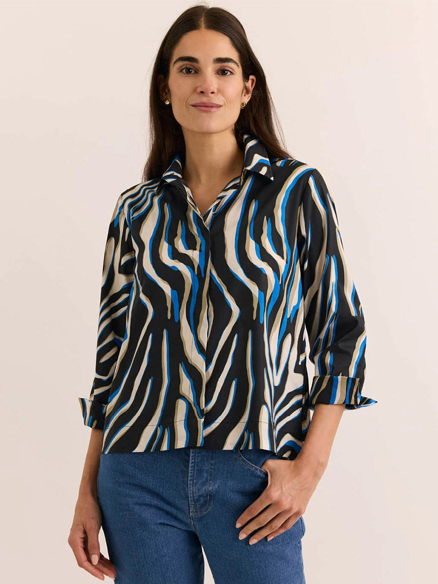 A woman wearing the Hinson Wu New Xena 3/4 Sleeve Shirt in Abstract Zebra—featuring black, white, and blue wavy stripes in exclusive prints on comfortable stretch fabric—is paired with blue jeans and posed against a plain light background.