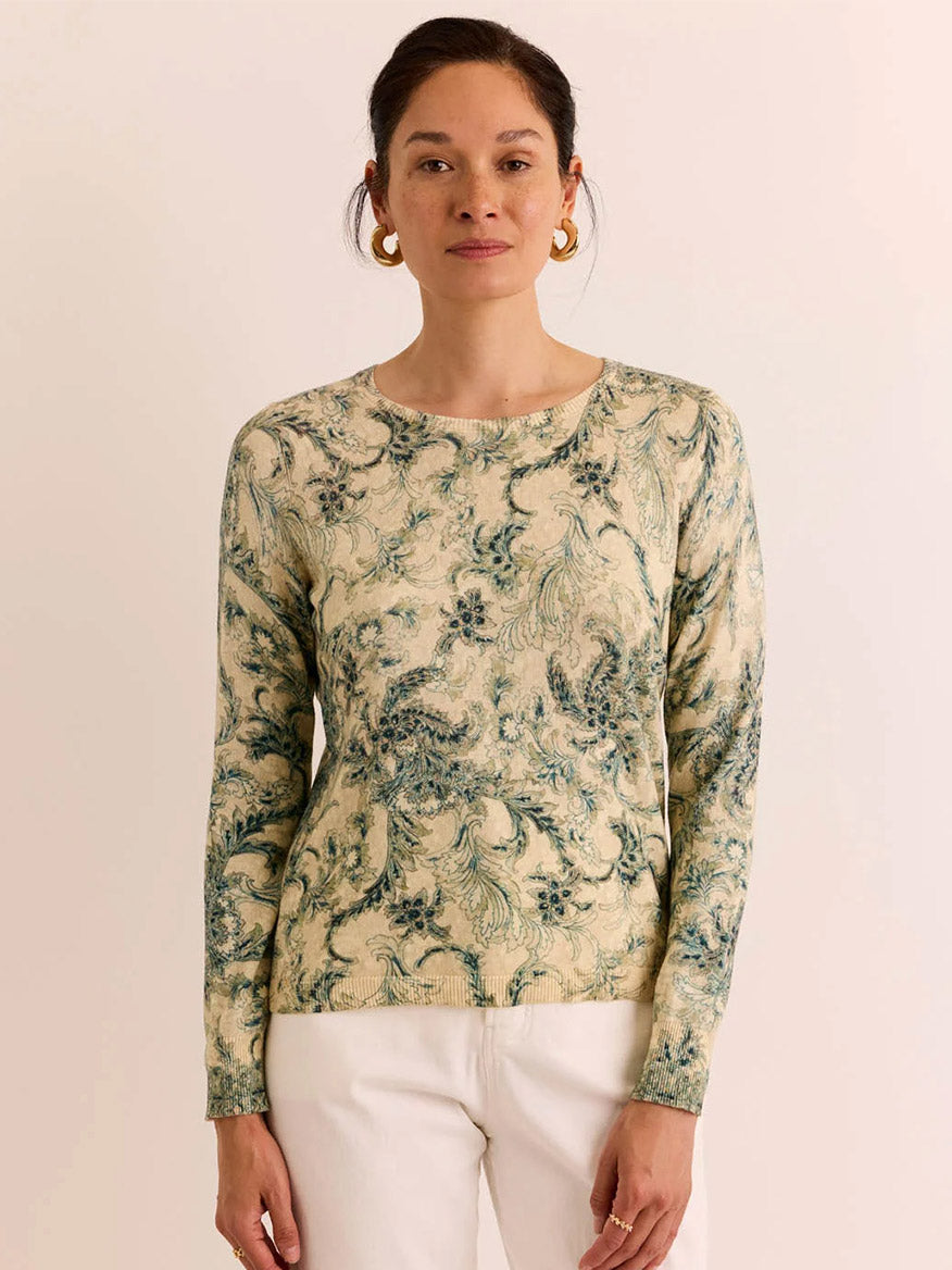 A woman with dark hair in a bun wears the Hinson Wu Phoebe Long Sleeve Fresco Print Sweater, featuring a blue and green leafy pattern on cream cotton, paired with white pants and standing against a plain light background. This comfortable sweater is the perfect wardrobe essential for any season.