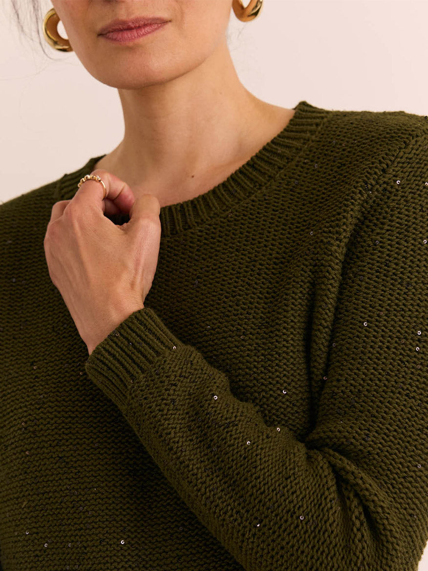 A woman wearing the Hinson Wu Queenie Long Sleeve Sequins Sweater in Oregano—a textured, olive green lightweight knit with subtle sequin embellishments—and gold hoop earrings, holding her left shoulder with her right hand. The breathable comfort of this sweater makes it ideal for year-round wear.