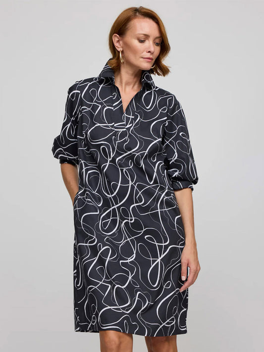 A woman stands with hands in pockets, wearing the Hinson Wu Vicky 3/4 Sleeve Ribbon Dress in Black/White featuring exclusive white abstract prints, set against a plain light gray background.