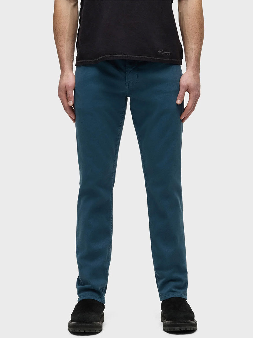 A man in Hudson Blake Slim Straight Jeans in Deep Dive and a black shirt stands against a plain background, presenting a sleek silhouette.