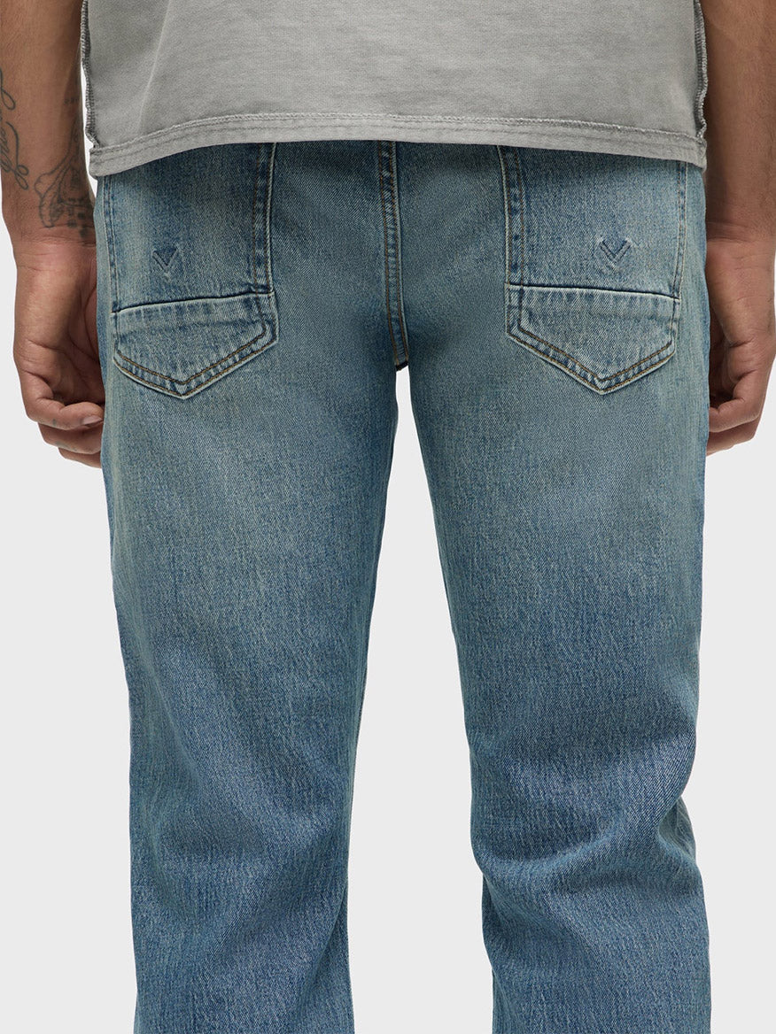 Back view of a person in Hudson Blake Slim Straight Jeans in Blue Cove, highlighting a medium indigo wash and comfort stretch denim. They are wearing a gray shirt with their arms at their sides.