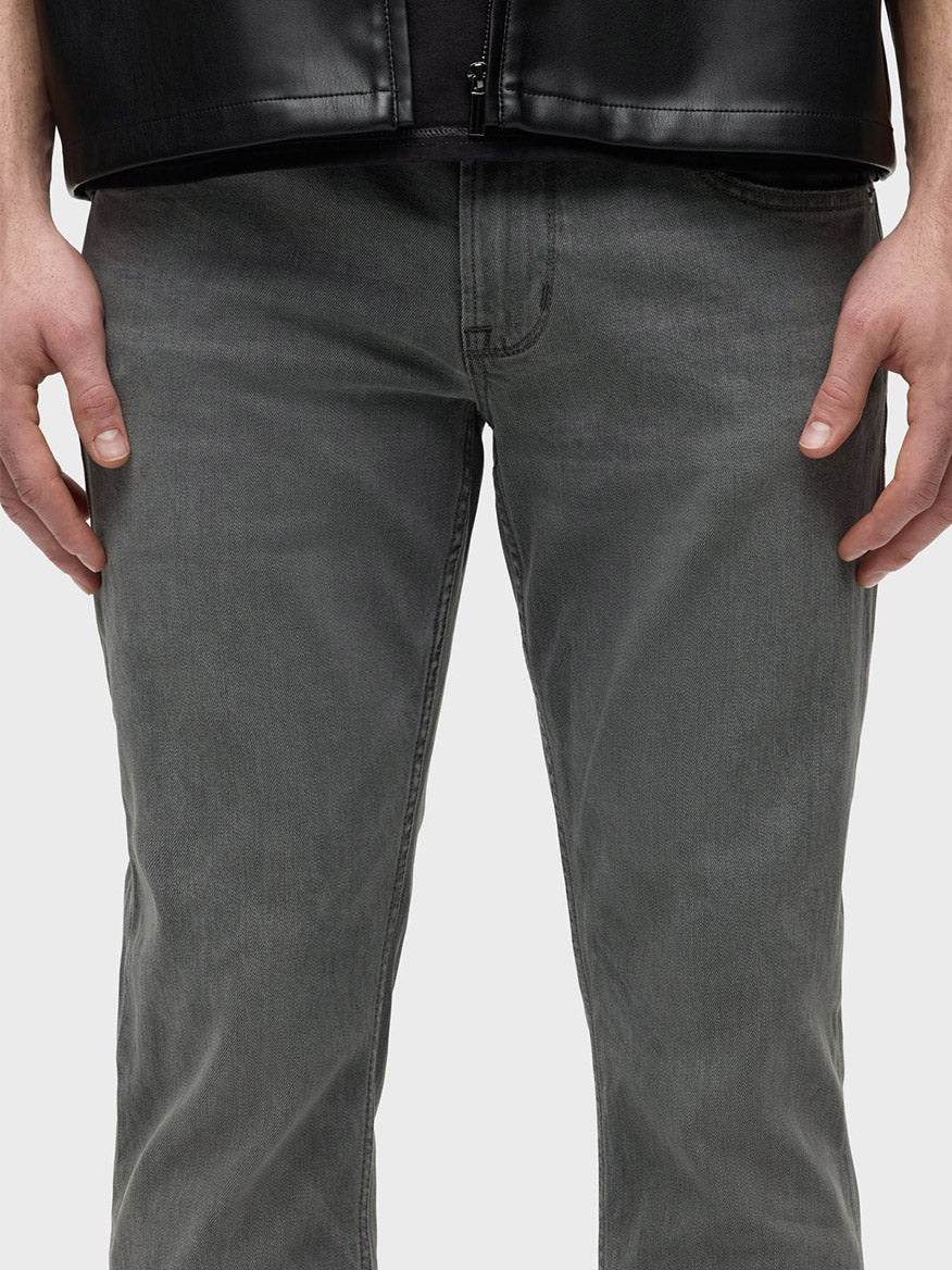 A person wearing Hudson Blake Slim Straight Jeans in Moon Ridge, which are gray with a tapered leg, and a black jacket, stands with hands at sides against a plain background.