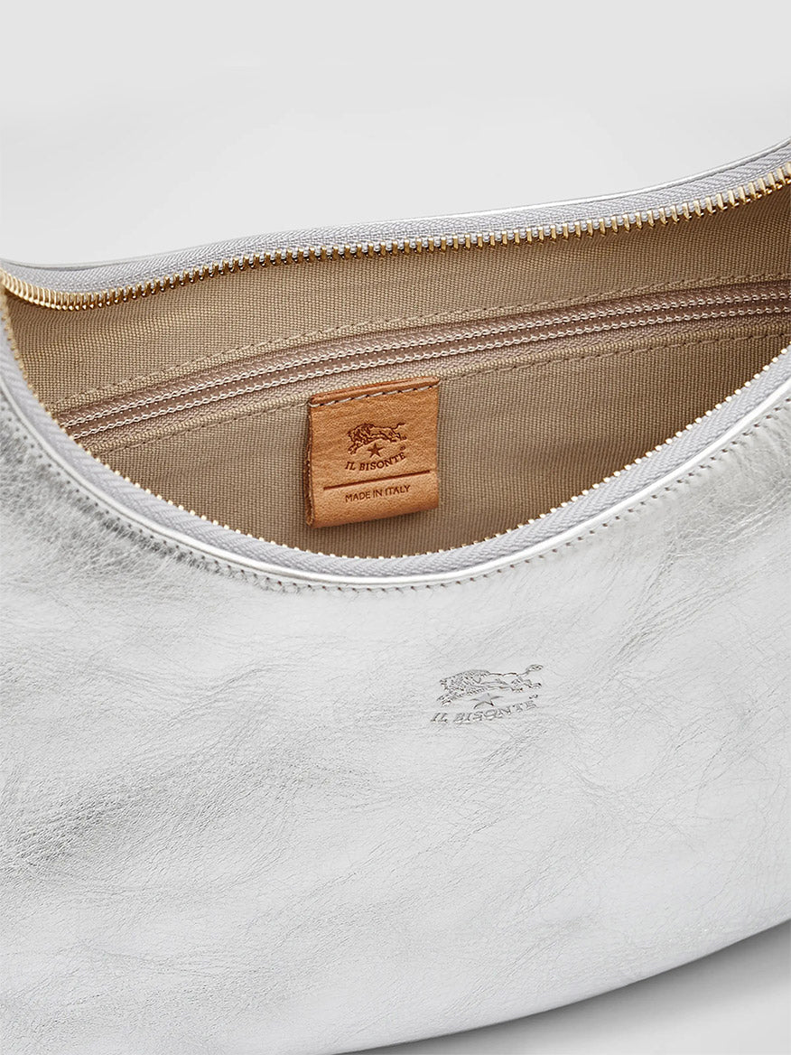 A close-up of an open white leather bag showing the beige interior and a tan leather label that reads "IL BISONTE MADE IN ITALY." Handmade in Tuscany, this Il Bisonte Belcanto Shoulder Bag in Metallic Silver features a gold zipper and an embossed logo on the exterior.