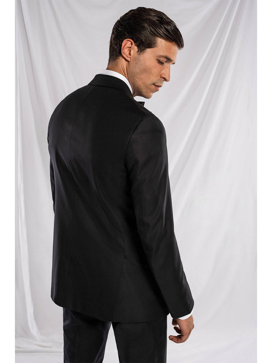 A man in an Isaia Sanita AquaSpyder Peak Lapel Tuxedo, white dress shirt, and black bow tie stands facing away in front of a white draped background, embodying the essence of Italian tradition with modern details.