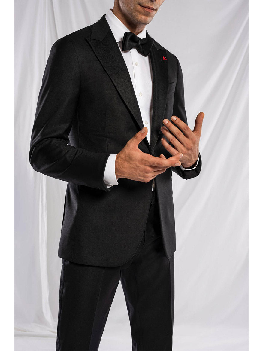 A man in a dark Isaia Sanita AquaSpyder Peak Lapel Tuxedo with a bow tie stands against a light background, showcasing a classic fit with modern details, gesturing with his right hand while his left hand rests at his side.