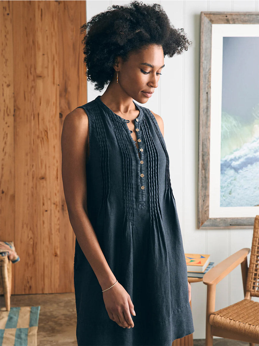 A woman in a black, lightweight fabric Faherty Brand Isha Basketweave Dress in Washed Black stands indoors, looking down thoughtfully, with a wooden wall and framed art in the background.