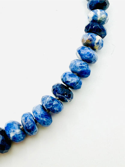 A close-up of the JMNYC Designs 18" Faceted Sodalite Rondelle Necklace showcases sparkling blue and dark navy beads arranged in a graceful curve on a white background—highlighting its elegant diamond clasp.