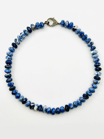 An 18-inch faceted sodalite rondelle necklace from JMNYC Designs, featuring blue and white stones and finished with a diamond clasp in sterling silver, arranged in a circular shape on a plain white background.