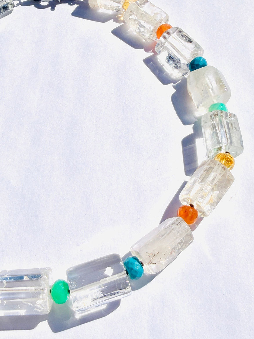 Introducing the JMNYC Designs 18" Crystal Quartz Cylinder Multi Stone Ball Necklace, a striking piece showcasing large clear rectangular crystal quartz cylinder beads paired with smaller round beads in vibrant turquoise, orange, and yellow hues. Presented on a white surface under bright light, this necklace is elegantly finished with a sterling silver clasp.