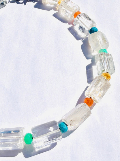 Introducing the JMNYC Designs 18" Crystal Quartz Cylinder Multi Stone Ball Necklace, a striking piece showcasing large clear rectangular crystal quartz cylinder beads paired with smaller round beads in vibrant turquoise, orange, and yellow hues. Presented on a white surface under bright light, this necklace is elegantly finished with a sterling silver clasp.