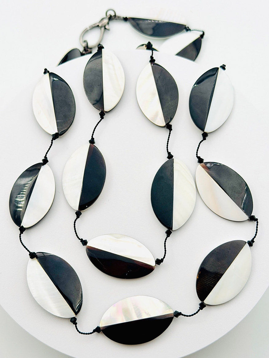 Introducing the JMNYC Designs 36" Oval Mother of Pearl and Black Tab Necklace—a bold statement piece showcasing oval beads split diagonally into black and mother of pearl halves. Each bead is artfully strung on a sleek black cord, creating a striking monochrome necklace.