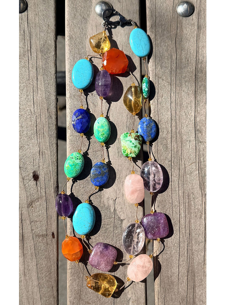 The JMNYC Designs 36" Vivid Multi Stone Necklace is displayed on a wooden surface, featuring large gemstones in vibrant shades of blue, green, purple, orange, and yellow. This eye-catching necklace boasts a stunning blend of colors, making it a truly unique accessory.