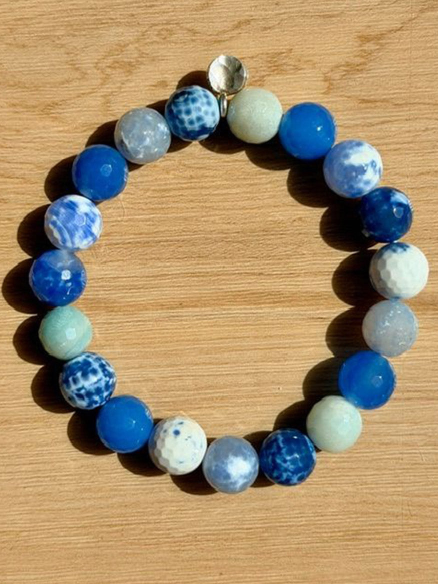 The JMNYC Designs Faceted Blue Agate, Mystic Aqua, Snow Peri, Denim Ball Bracelet—with its alternating blue and white marbled beads—lies arranged in a circle on a wooden surface.