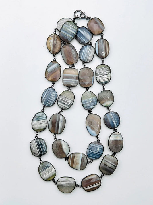 The JMNYC Designs Large Blue Lace Agate Necklace With Rhodium Bezel & Clasp 36" features double layers of flat, irregular oval Blue Lace Agate stones with gray, brown, and white stripes linked by rhodium-plated metal.