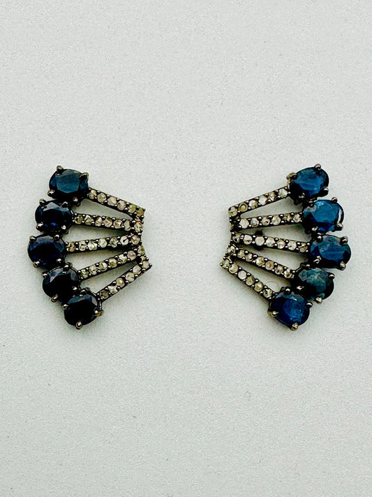 The JMNYC Designs Blue Sapphire Diamond Sterling Silver Post Earring features three rows of round blue sapphires and small clear stones in a fan-like, geometric design on a light sterling silver background.
