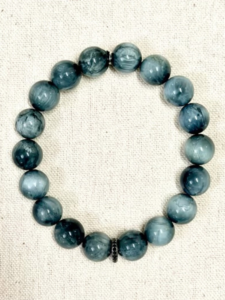 A JMNYC Designs Eagle Eye Stretch Bracelet with Sterling Silver Black Spinel Rondelle, featuring round, marbled blue-gray stones, is arranged in a circle on a beige fabric background.