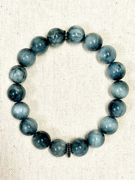 A JMNYC Designs Eagle Eye Stretch Bracelet with Sterling Silver Black Spinel Rondelle, featuring round, marbled blue-gray stones, is arranged in a circle on a beige fabric background.