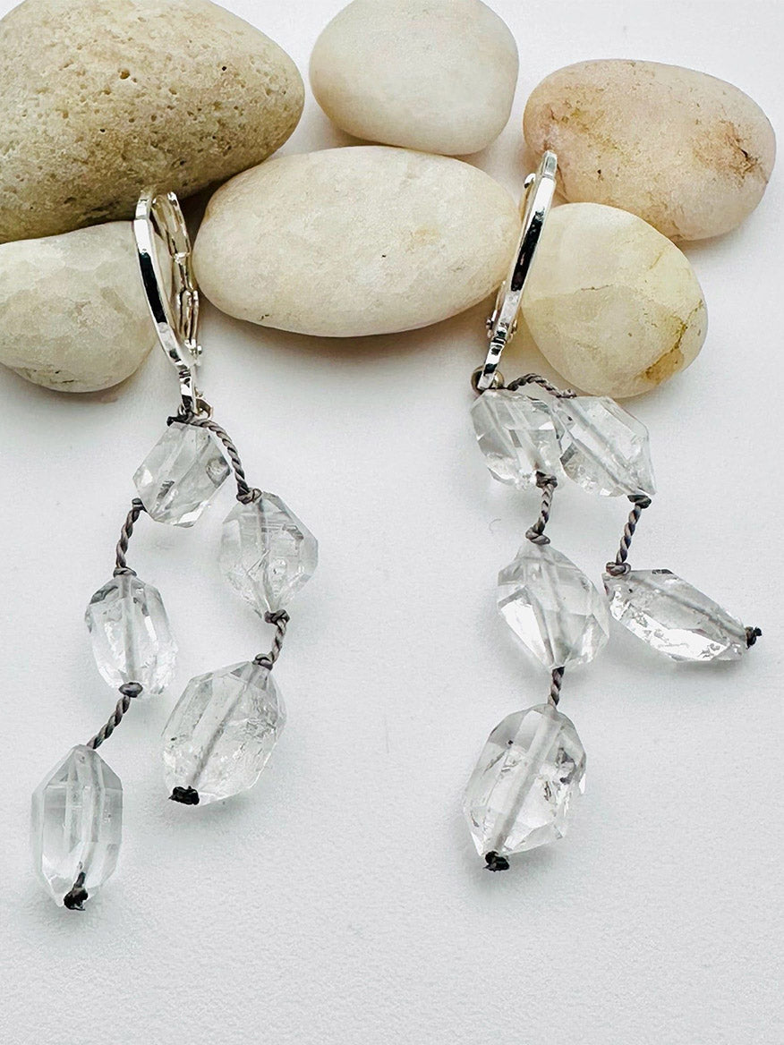 Introducing the JMNYC Designs Flat AAA Faceted Clear Herkimer Diamond Earring on Sterling Silver Huggie, 2": This limited edition pair features sparkling, faceted Herkimer diamond beads strung on gray cord and suspended from sleek sterling silver huggie hoops. Displayed on a white surface alongside smooth white stones, these earrings offer a contemporary twist to classic elegance.