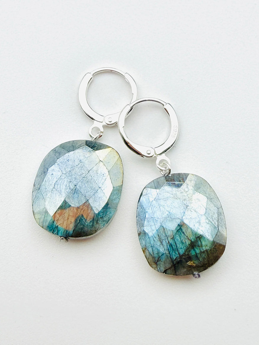 The JMNYC Designs Flat Faceted Mystic Labradorite Earrings feature large, blue and green iridescent labradorite drops set on sterling silver huggie lever-backs, shown against a white background.