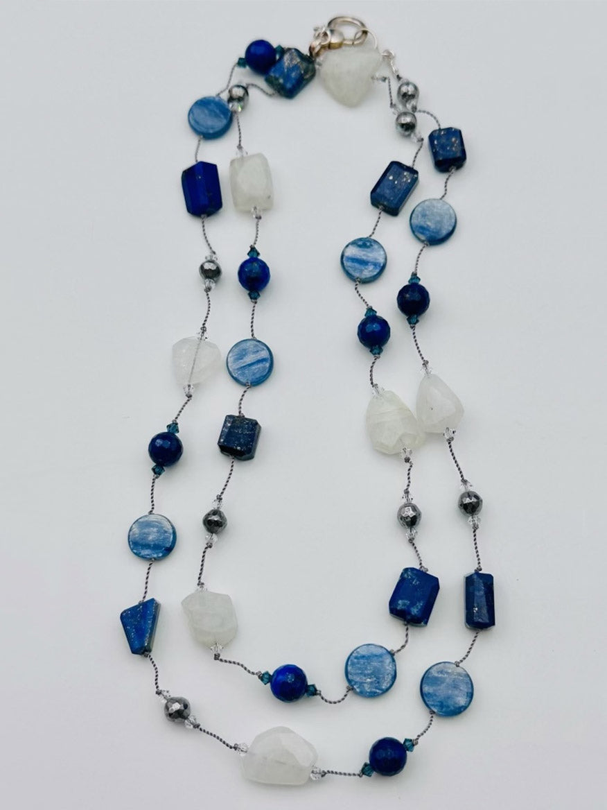 The JMNYC Designs Kyanite, Lapis, Rainbow Moonstone & Pyrite Long Necklace showcases an elegant blend of blue kyanite, deep lapis, shimmering rainbow moonstone, and sparkling pyrite beads in a single strand. Artfully arranged with stones of various shapes and sizes on a white background, this piece radiates serene sophistication.