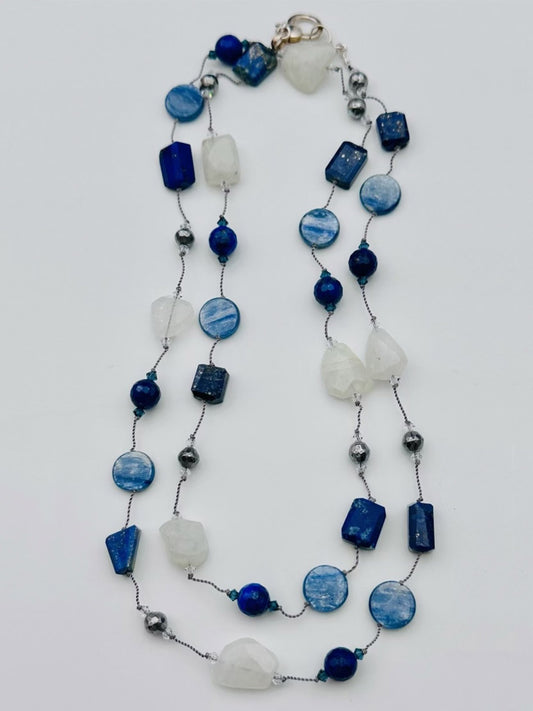 The JMNYC Designs Kyanite, Lapis, Rainbow Moonstone & Pyrite Long Necklace showcases an elegant blend of blue kyanite, deep lapis, shimmering rainbow moonstone, and sparkling pyrite beads in a single strand. Artfully arranged with stones of various shapes and sizes on a white background, this piece radiates serene sophistication.