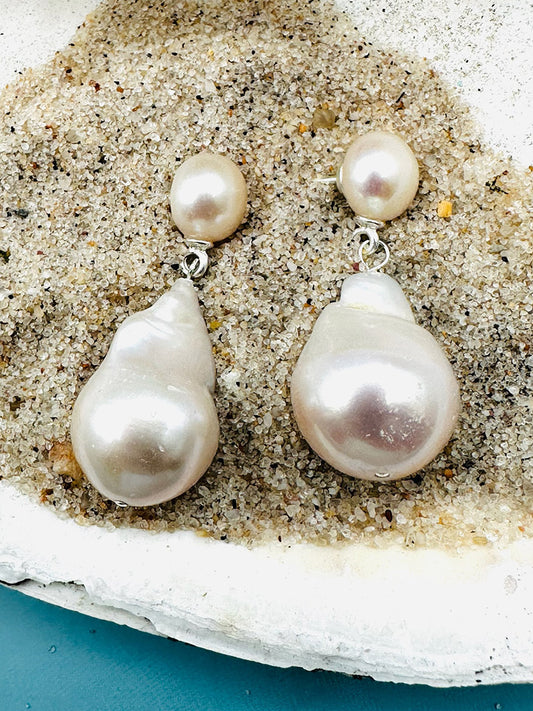 A pair of JMNYC Designs Medium White Freshwater Baroque Pearl Earrings with Freshwater Pearl Post displayed on coarse sand inside a white shell.