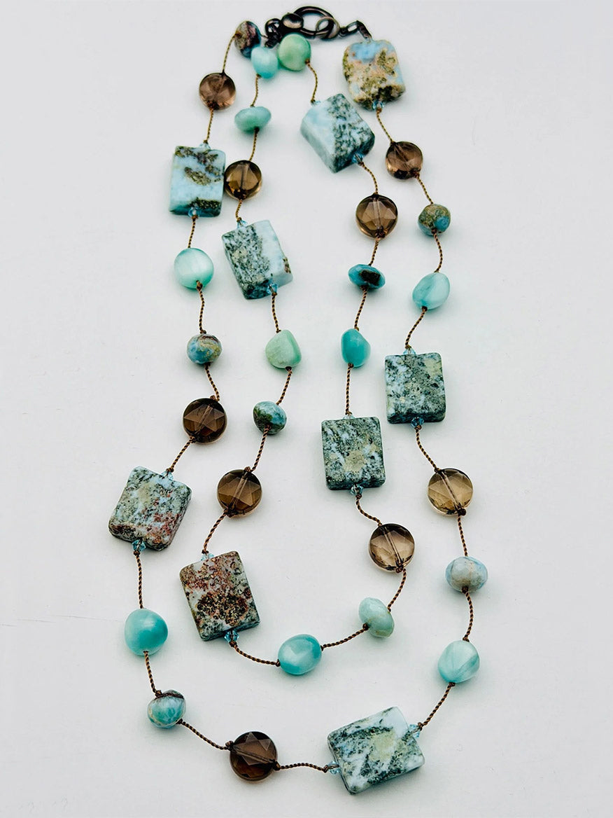 The JMNYC Designs Organic & Smooth Larimar & Smokey Quartz Combo Necklace showcases an elegant mix of rectangular Larimar green stones, round Smokey Quartz brown beads, and small turquoise stones delicately strung on a fine string, bringing together earthy tones reminiscent of nature’s beauty, displayed gracefully against a white background.