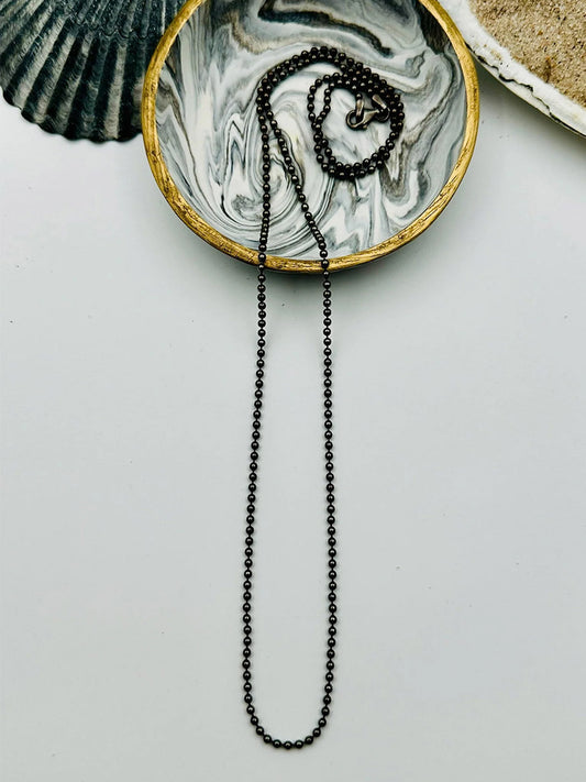 The JMNYC Designs Sterling Silver Rhodium Ball Chain Necklace 24" is shown on a round, marbled dish with a gold rim, beside seashells on a white surface.