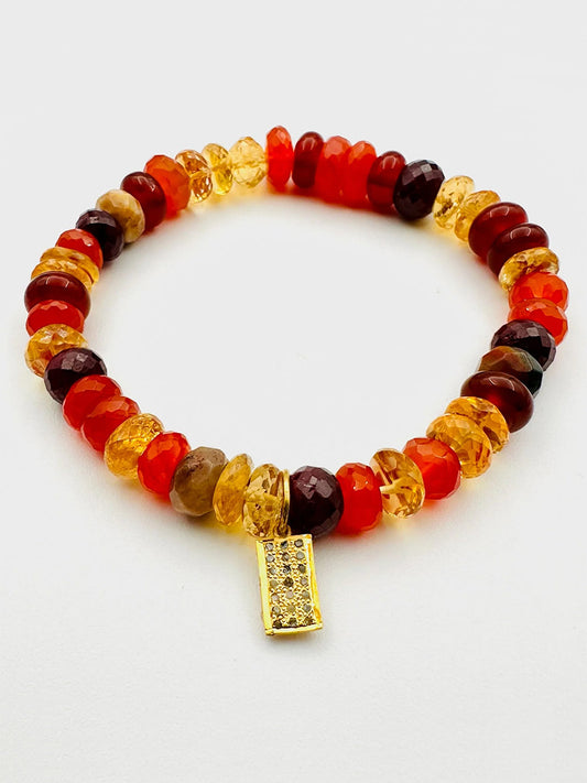 The JMNYC Designs Ruby, Carnelian & Citrine Stretch Bracelet features red, orange, and yellow faceted stones with a gold dog tag charm accented with rhinestones. Shown on a white background.