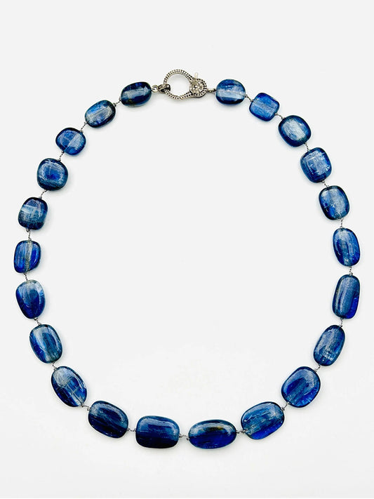 The JMNYC Designs Smooth Oval Kyanite Nugget Necklace, showcasing lustrous blue oval stones and finished with a Sterling Silver Diamond Clasp, is elegantly displayed in a circle on a plain white background.