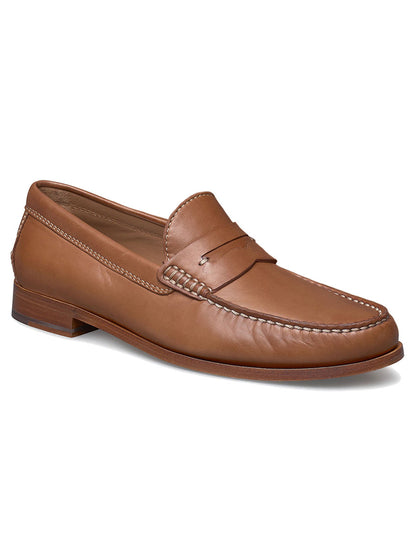 The J & M Collection Baldwin Penny in Cognac Sheepskin is a brown leather loafer with moccasin construction, featuring a low heel, penny slot detail, and white stitching around the edges.