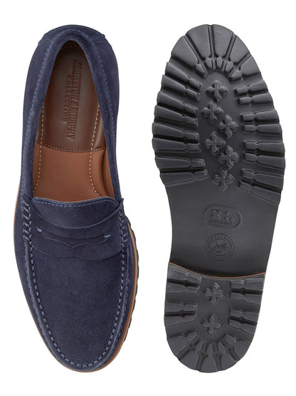 The J & M Collection Baldwin Lug Penny in Navy English Suede loafers are shown in side and sole views, featuring a leather lining and an EVA lug outsole with a textured pattern of symbols and lettering. A brown interior is visible.