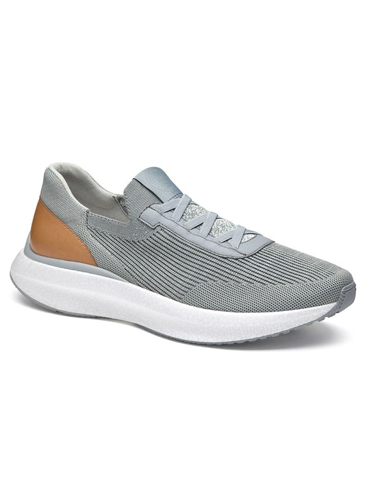 The J & M Collection Briley Knit Jogger in Grey Knit is a single grey sneaker featuring a white sole and a tan heel accent. It boasts knit uppers with a mesh texture, elastic laces, and a memory-foam footbed for ultimate comfort.