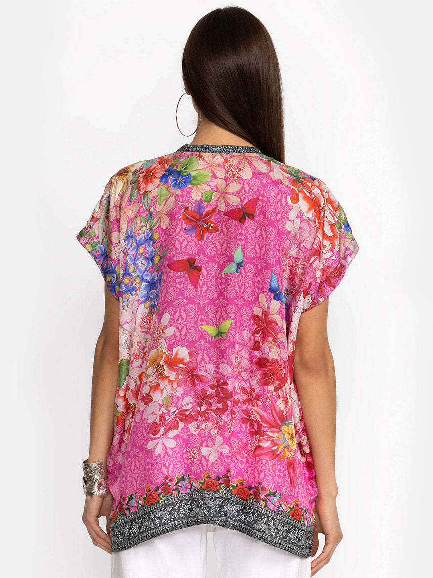Replace "Bouquet Frame Parade Blouse" with "Johnny Was Bouquet Frame Parade Blouse in Multi"
Sentence: Rear view of a woman wearing a vibrant floral printed Johnny Was Bouquet Frame Parade Blouse in Multi with pink, orange, and blue colors, accessorized with silver bracelets and a hoop earring.