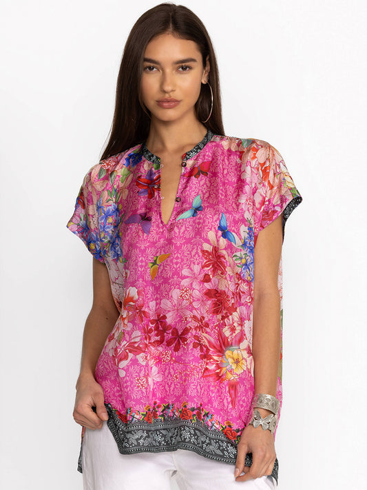 A woman modeling a vibrant floral print Johnny Was Bouquet Frame Parade Blouse in Multi with draped dolman sleeves, standing against a white background.
