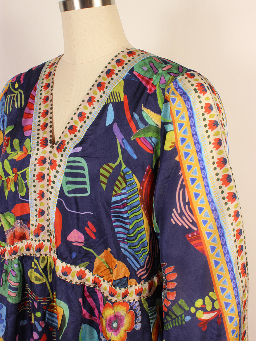 A mannequin displays the Johnny Was Saskia Midi Dress in Farout Scarf Print, a colorful silk creation featuring a vibrant pattern of abstract shapes and flowers on a dark background, ornate trim along the neckline, and flowing kimono sleeves.