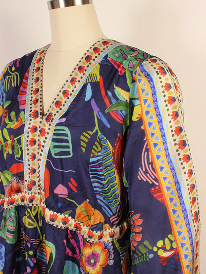 A mannequin displays the Johnny Was Saskia Midi Dress in Farout Scarf Print, a colorful silk creation featuring a vibrant pattern of abstract shapes and flowers on a dark background, ornate trim along the neckline, and flowing kimono sleeves.