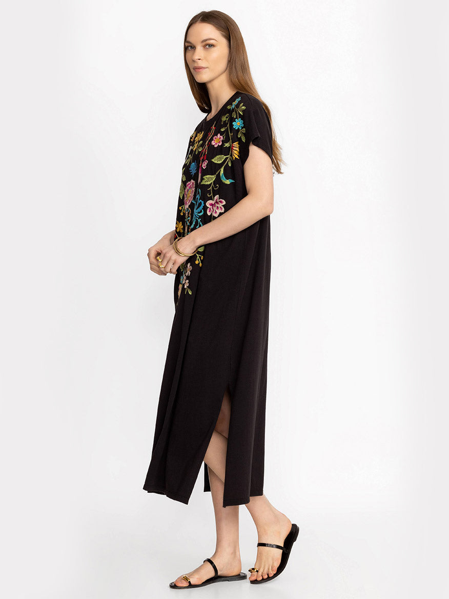 A woman standing in a profile view, wearing a Johnny Was Sheri Relaxed Knit Dress in Black with colorful placement embroidery, paired with black sandals.
