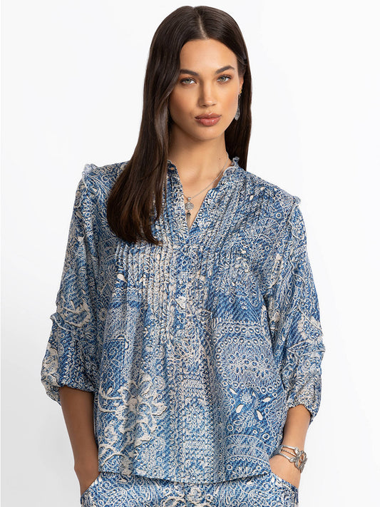 A woman stands in a Johnny Was Zeeland Blouse featuring a denim print, adorned with luxurious intricate detailing, her expression neutral against a plain background.