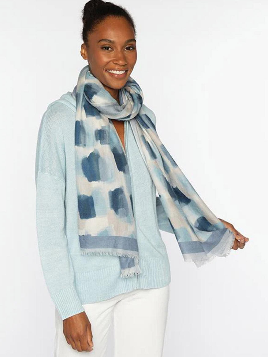 A woman smiles against a plain white background, wearing a light blue sweater, white pants, and the Kinross Brushstroke Print Scarf in Ink Multi.